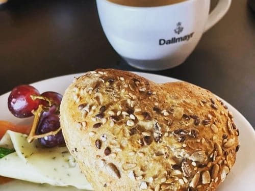 Herzförmiges Mehrkornbrötchen mit Käse, Trauben und einer Tasse Kaffee.