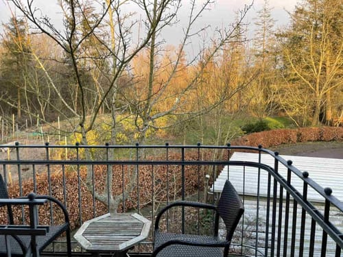 Ein kleiner Balkon mit zwei Stühlen und einem Tisch, mit Blick auf einen herbstlichen Garten und kahlen Bäumen.