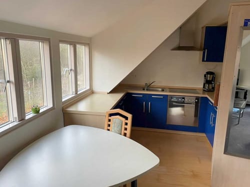 Helle Dachgeschossküche mit blauen Schränken, Esstisch und großen Fenstern mit Blick ins Grüne.