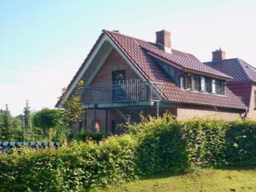 Ein modernes Einfamilienhaus mit rotem Ziegeldach und Balkon, umgeben von einem üppigen Garten und Hecken.