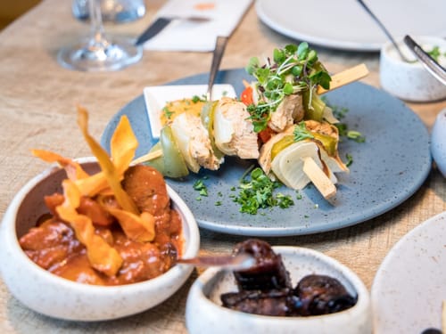 Eine Auswahl an köstlichen Tapas auf einem Holztisch, darunter Spieße mit Hähnchen und Gemüse.