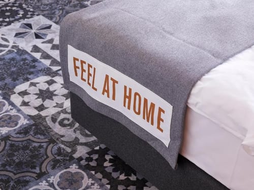Gepflegtes Bett mit grauer Tagesdecke und der Aufschrift "Feel at Home" auf einem gemusterten Teppichboden.