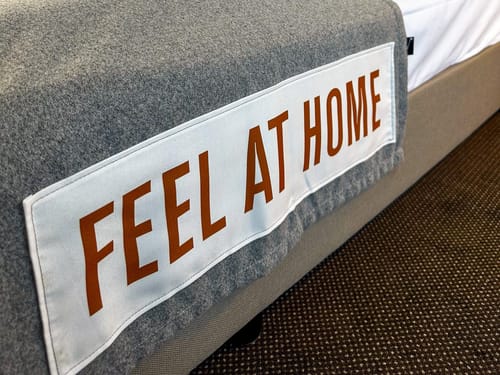 Gemütliches Bett mit grauer Decke und Aufschrift "Feel at Home"