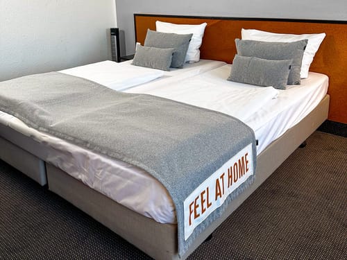 Gemütliches Doppelbett in einem modernen Hotelzimmer mit der Aufschrift "Feel at Home" auf der Tagesdecke.