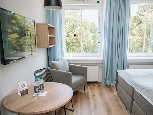 Modern eingerichtetes Hotelzimmer mit großem Fenster, grauem Sessel und rundem Holztisch