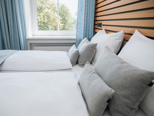 Modernes Doppelbett mit weicher Bettwäsche und grauen Kissen vor einem großen Fenster mit Blick auf grüne Bäume