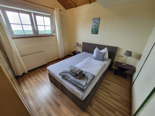 Gemütliches Schlafzimmer mit Doppelbett, Fenstern mit Vorhängen, Holzboden und neutralen Farben.