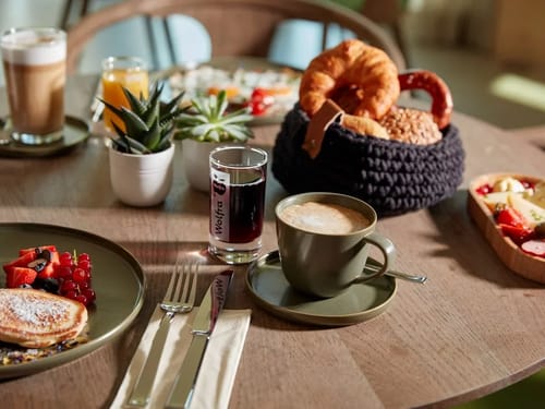 Gemütliches Frühstück mit Kaffee, frischen Brötchen, Croissants, frischem Obst und Säften auf einem Tisch.