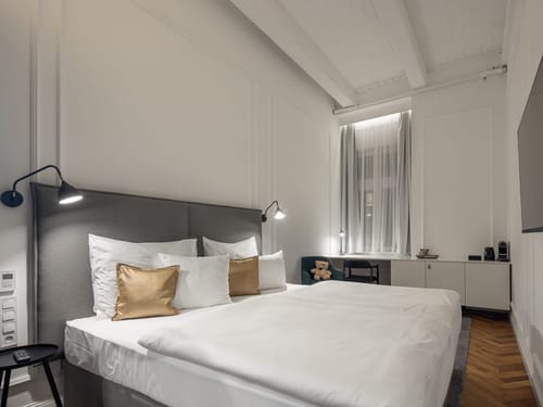 Helles und stilvoll eingerichtetes Hotelzimmer mit einem großen Doppelbett, weißer Bettwäsche und goldenen Akzentkissen vor einem gepolsterten grauen Kopfteil. Wandleuchten in Marineblau sorgen für warmes Ambientlicht, während ein Schreibtisch mit Stuhl und eine Kaffeemaschine im Hintergrund für Komfort sorgen