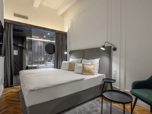 Stilvoll eingerichtetes Hotelzimmer mit einem großzügigen Doppelbett, grauem Polsterkopfteil und weißer Bettwäsche mit goldenen Zierkissen.