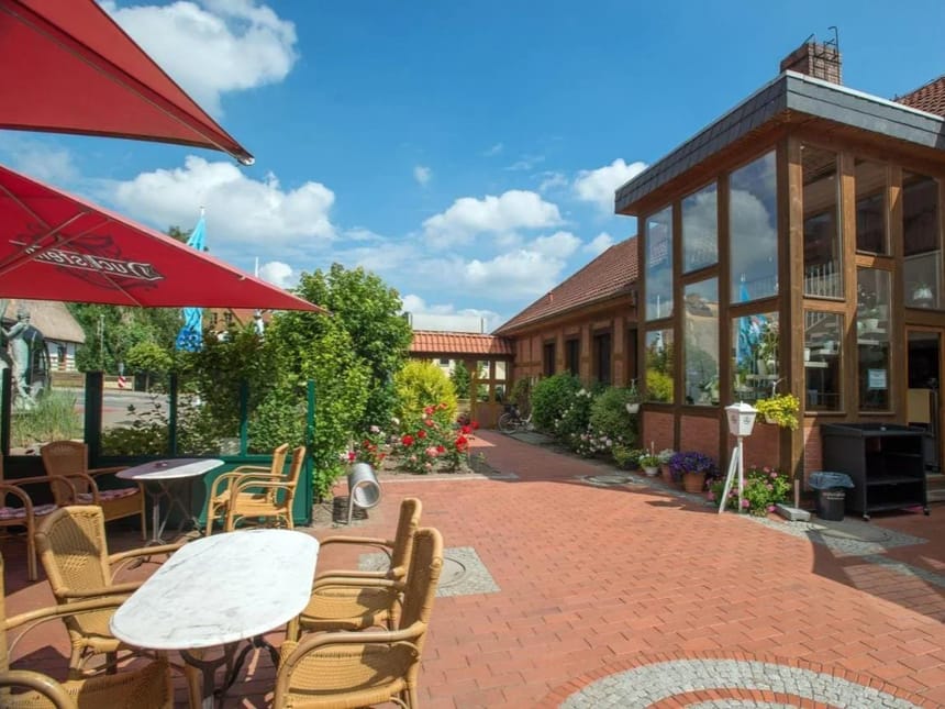 Sonnige Terrasse eines Cafés mit gemauertem Boden, Holzmöbeln und umgeben von Pflanzen.