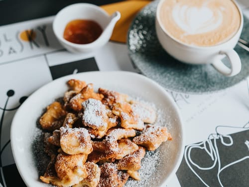 Eine Portion Kaiserschmarrn auf einem Teller mit Puderzucker bestäubt, serviert mit einer Tasse Cappuccino und einem Schälchen Marmelade.