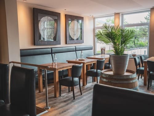 Modernes Restaurant-Interieur mit eleganten Holztischen, schwarzen Stühlen, großen Pflanzen und stilvollen Wanddekorationen.