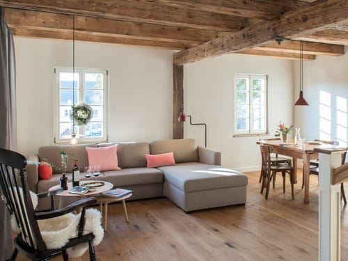 Gemütliches Wohnzimmer mit Holzbalkendecke, einem großen grauen Sofa, Esstisch und hölzernem Schaukelstuhl