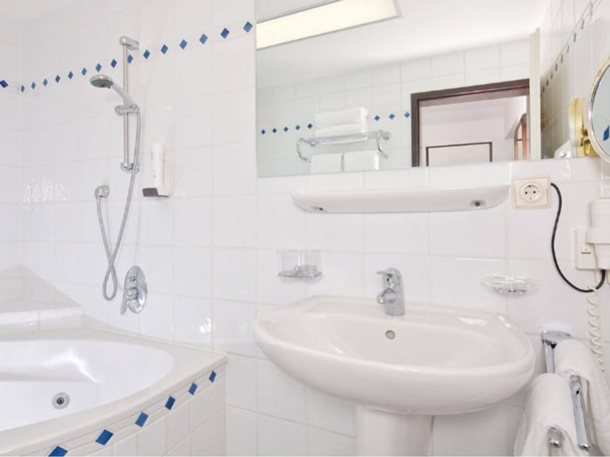 Gepflegtes Hotelbadezimmer mit Badewanne, Dusche, Waschbecken und Spiegel in klassisch-weißem Design mit blauen Akzenten