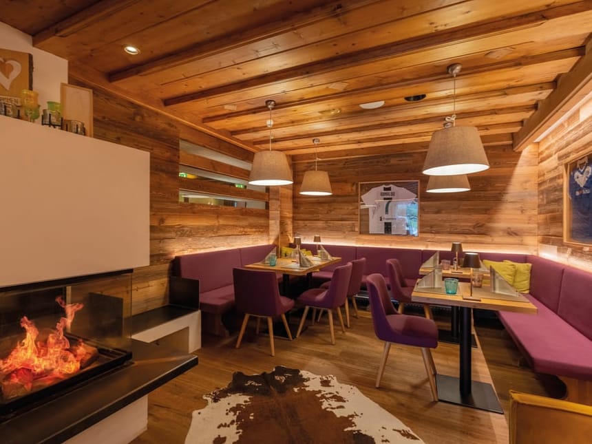 Gemütlicher Gastraum mit Kamin, rustikaler Holzverkleidung und lila Sitzmöbeln in einem alpinen Restaurant