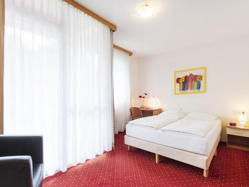 Modernes Hotelzimmer mit Doppelbett, roten Teppichboden und großen Fenstern mit Vorhängen.