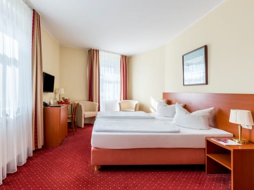 Hell eingerichtetes Hotelzimmer mit Doppelbett und rotem Teppichboden