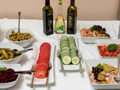 Verschiedene frische Salate und Beilagen auf einem Tisch, darunter Oliven, Tomaten, Gurken, Essiggurken und Öl.