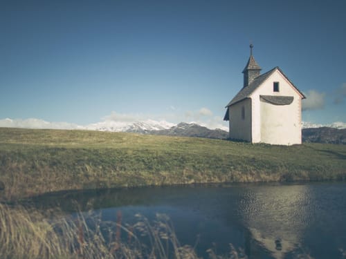 Kleine Kapelle auf einer Wiese vor einer Bergkulisse mit Wolken und blauem Himmel.