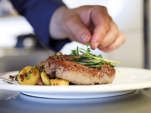 Professioneller Koch beim Anrichten eines Steaks mit Kräutern und Bratkartoffeln