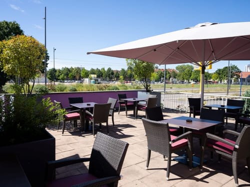 Sonnige Außenterrasse eines Cafés mit Tischen und Stühlen aus Rattan unter einem großen Sonnenschirm.